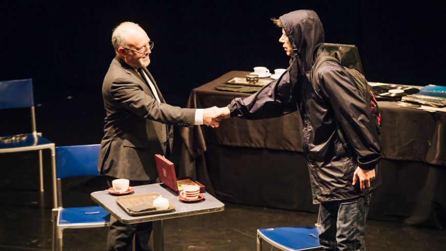 Two men greet each other with a handshake. One is dressed in a suit, the other in a rain coat with a hood.