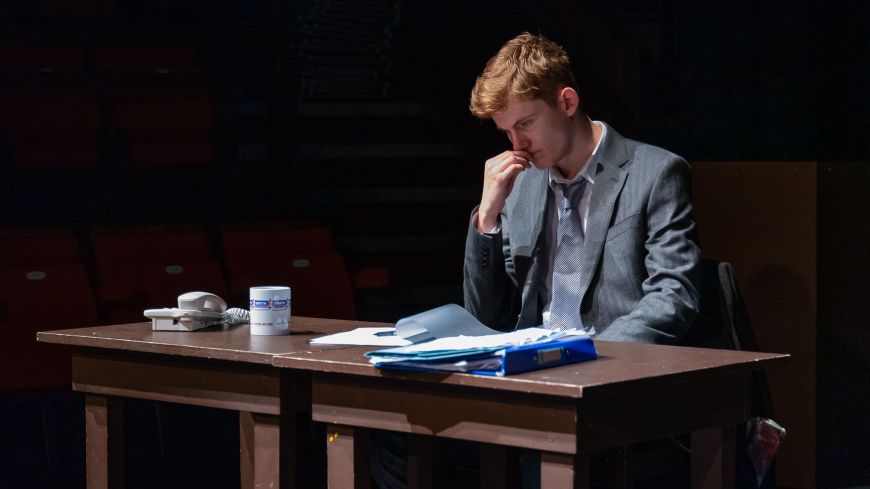 The lead actor as The Lawyer, sits at a table looking at papers, with a thoughtful look on his face.