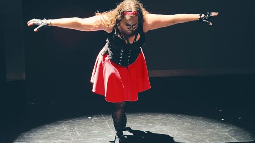 Performer Torrey Shine, dressed in black bustier, pink skirt, looks down with her arms outstretched, lit in a circle, but surrounded by darkness