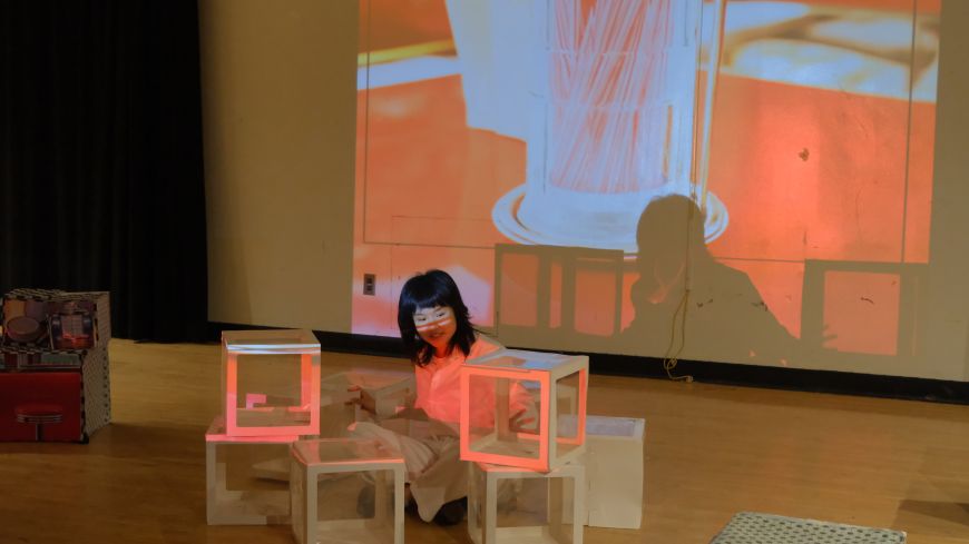 Performer Xiner Lan sits on the floor in front of white and orange boxes, with a projection screen in the background, shown with an orange wash