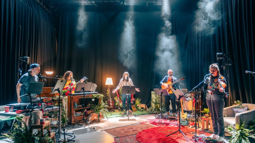 Five performers are light in a smoky atmosphere, with greenery, in a half circle