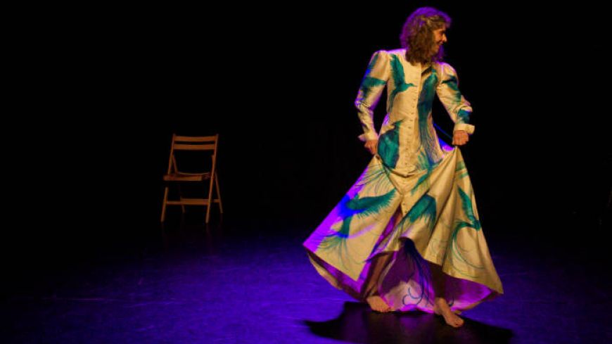 Performer Maria MacDonell starts, highlighted in a dark background, in a long glamorous cream coloured robe that has large bright blue peacocks on it.