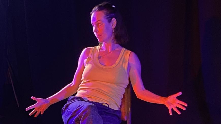 Performer Ambrosine Davies sits in a chair, with her arms extended, in dark light with a spotlight. She wears dark blue jogging trousers and a lime green vest.