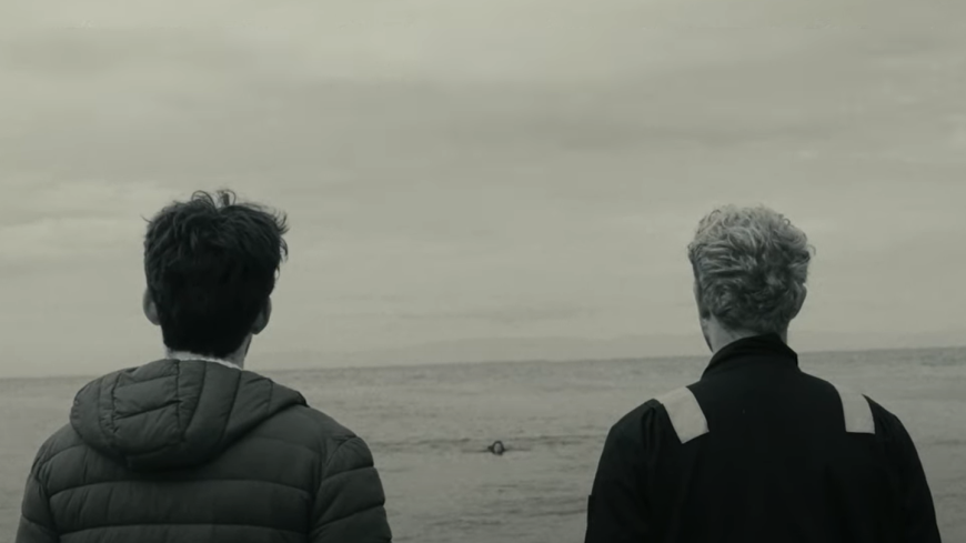 Two young men stand and look out at the wide open sea. The day seems grey and gloomy.