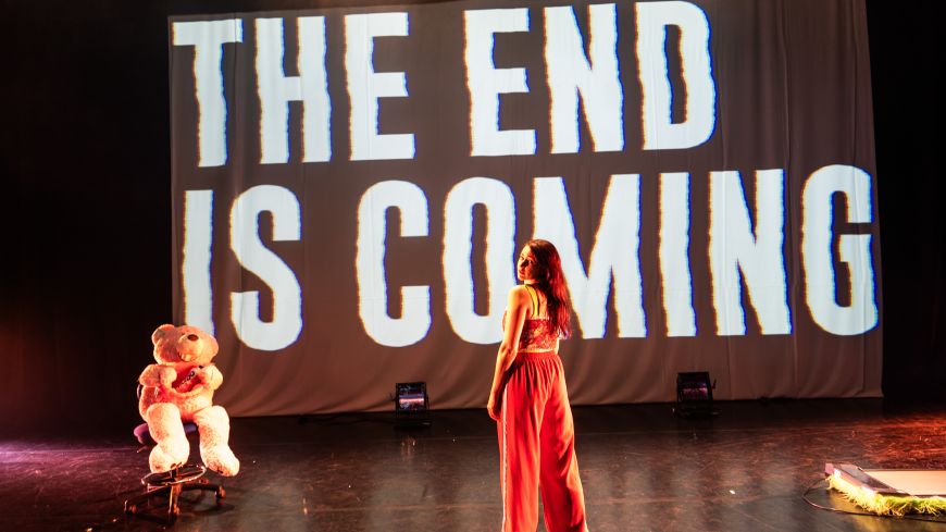 Screen with the words 'the end is coming' angled across it. Performance artist Louise Orwin stands in front of it with her back to the audience, wearing a red strappy top and red tracksuit bottoms. A giant teddy bear is seated to the left of the screen.