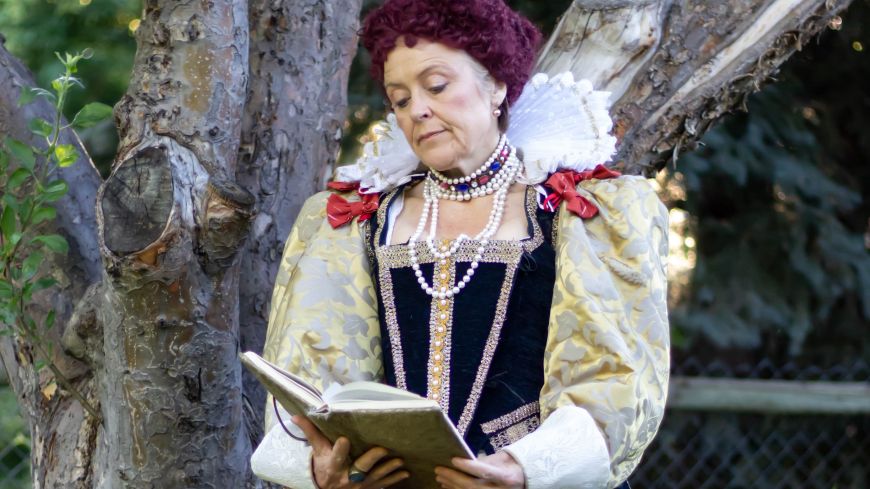 Elizabeth in her wig and full costume, looking at a book with her head tilted.