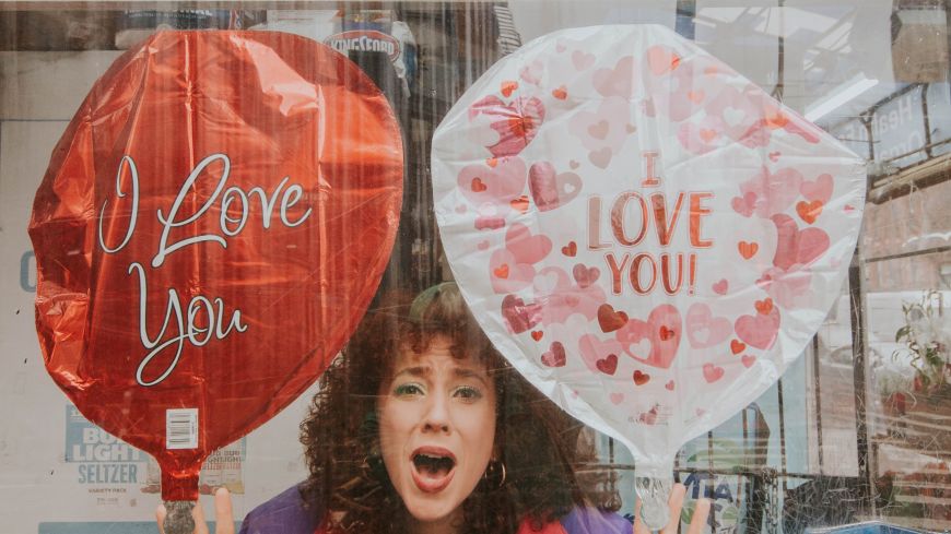 Writer/Performer Gianna Milici holds two balloons, one red, one pink. She wears a long green scarf around her neck.