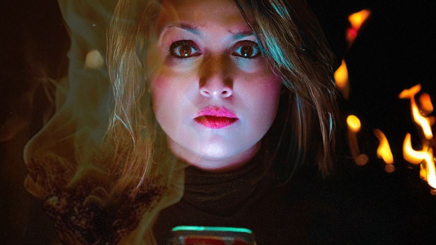 Woman looks directly at the screen. Darkness is behind with some fire. Fierce look.