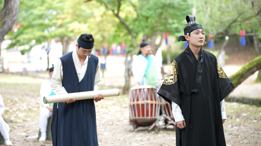 A courtier stands to the left of the King, with the Queen in the background, pictured in a green leafy area.