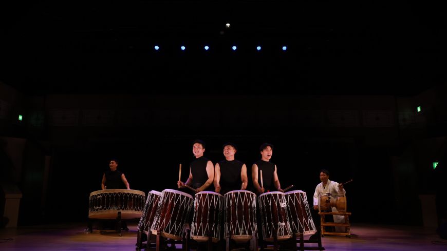 Drummers are pictured with their drums, in front of a dark background.