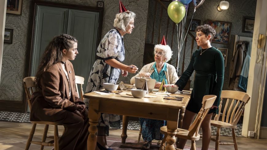 The four actors in the play are pictured round a kitchen table, all looking at each other, balloons in the background.