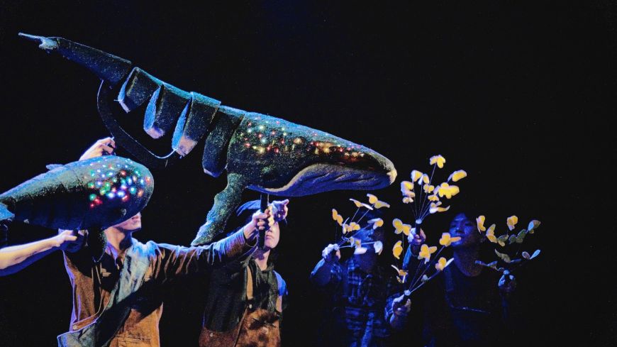 Whale puppet moves, held by puppeteers. Dark background, whale is lit up with lights showing on head.
