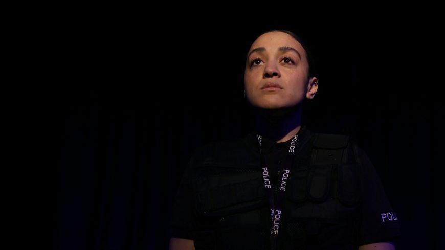 PC Irie looks out into the world, a serious look on her face. There is darkness around her but she is in the spotlight.