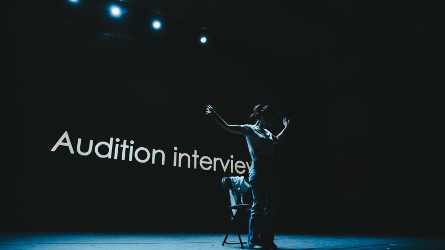 Hyojin Park stands in front of a screen on which the words 'Audition Interview' appear. The background is dark with the performer standing with arms outstretched in silhouette.