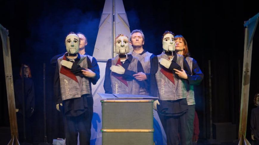 Performers on stage with the puppet masks, stand in the centre, large item at the back behind them