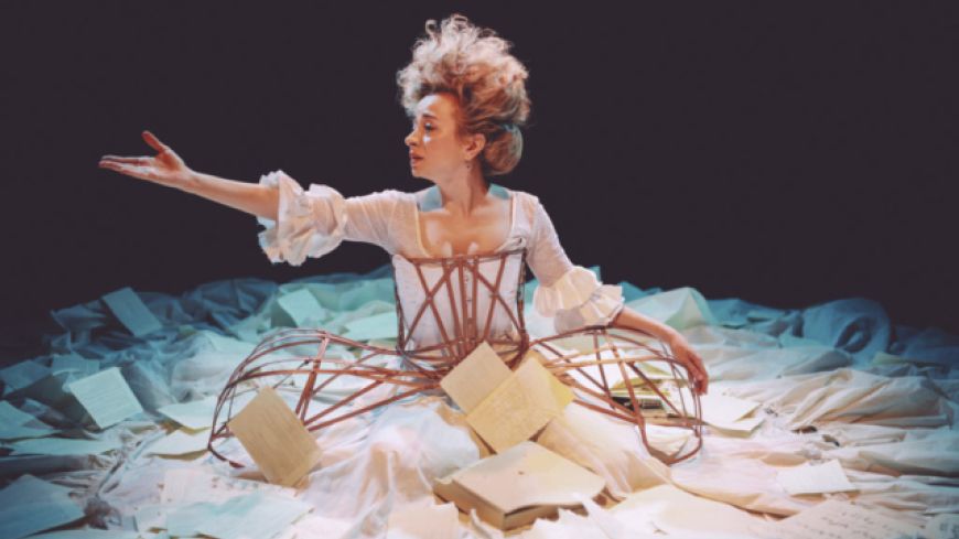 A woman sits on the ground, surrounded by a big white dress. Her hair is piled high and messy and one arm is outstretched.