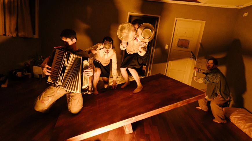 A musican plays; 3 other actors hide behind objects in a dark, shadowy room.