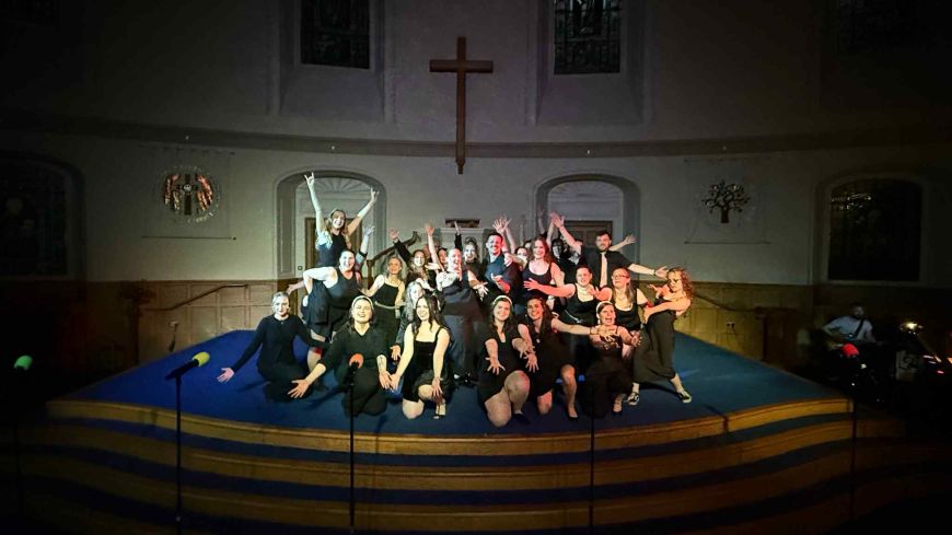 Large cast photo pose with arms out, black background, all seen in the spotlight. Church background shown.