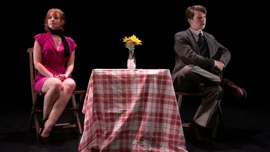 The two actors are pictured on stage, the female Comrade to the left, the male Agent on the right, with a dinner table between them, covered in a red and white checked table cloth.