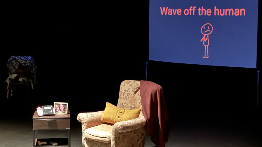 A screen shows in the background, an armchair and small bedside table to the side. The lighting is dark, with the furniture lit.