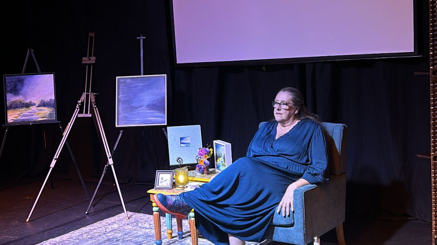 Performer/playwright Patricia McGrath sits next to a screen. There is dark blue light and paintings to the side. Everything is awash with shades of blue.