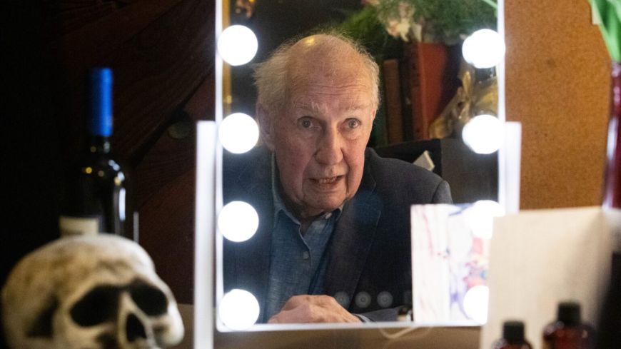 Nick Ullett, the actor and writer, is reflected in a dressing room mirror, lights around it, and a skull to his left.