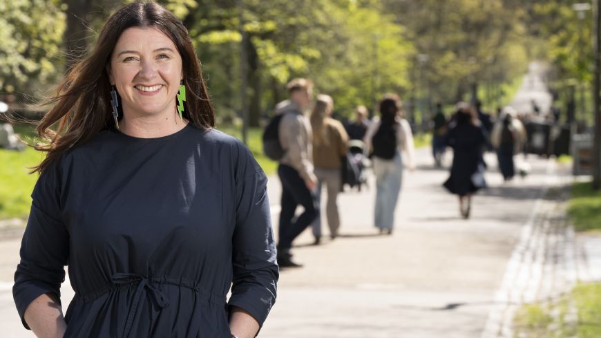 Book Festival director, Jenny Niven, wearing a black summer dress, smiles to the camera on Middle Meadow Walk. In the background, are walkers passing up the hill.