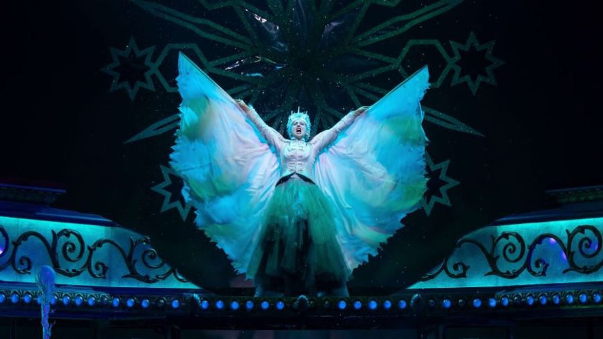 Claire Dargo as The Snow Queen, standing with her arms outstretched, reaching up to the sky. Black background with Snow Queen lit up in icy blue white.