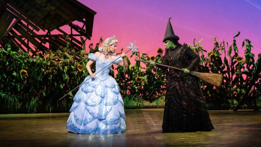 Sarah O'Connor and Laura Pick (Glinda and Elphaba) face off with brooms. Glinda wears a sparkly blue ballgown, Elphaba her traditional witch's outfit and has her green body colouring. 