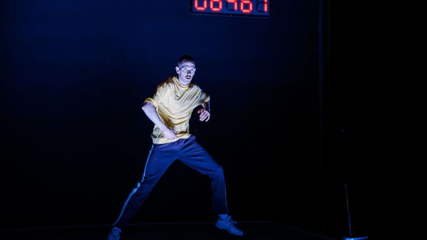 A male dancer is lit in the centre of a dark space with a screen showing numbers in red in the background