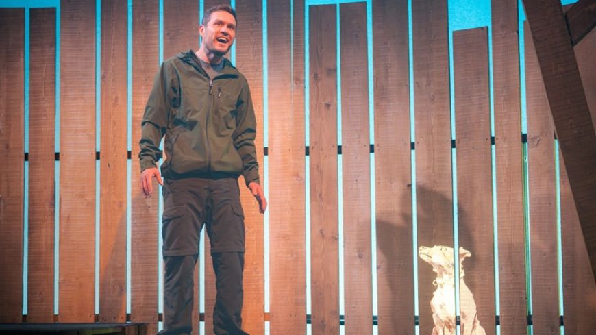 Jordan Young as Ronnie looking out and smiling with a fence behind him projecting an illustration of a small terrier dog.