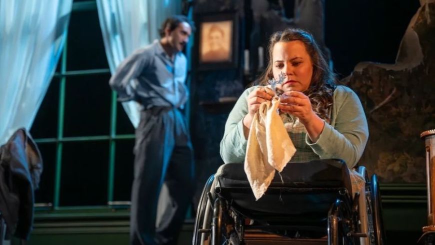 Amy Conachan as Laura sits polishing a glass figure as Christopher Jordan-Marshall as Tom hovers in the background arguing offstage with their mother.