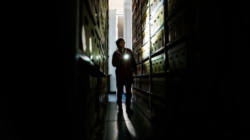 Danny Robins. strides between shelving, covered in box files, moody lighting in black and white