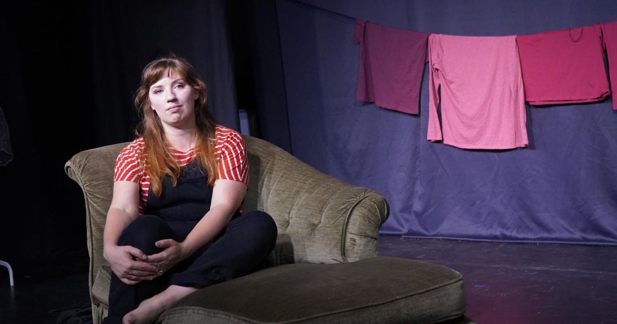 Performer sitting on a chaise longue, looking out, deep in thought. Background is dark, lit to focus on the performer and her laundry hanging behind (visual red flags).