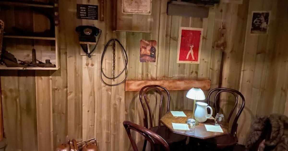A darkened room with a table, chairs, and wooden walls. Teapot and glasses on the table.