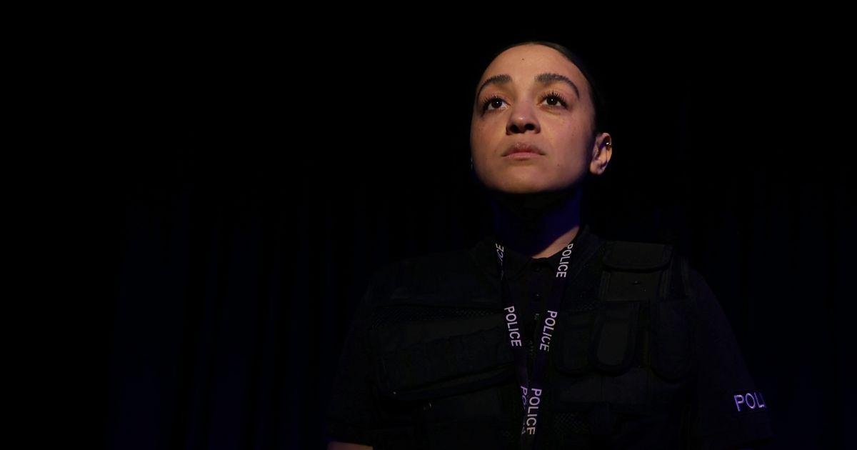 PC Irie looks out into the world, a serious look on her face. There is darkness around her but she is in the spotlight.
