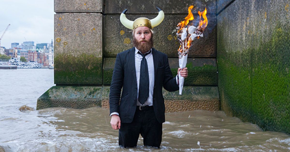 James Rowland, dressed in a suit with a viking helmet on his head, stands in the river