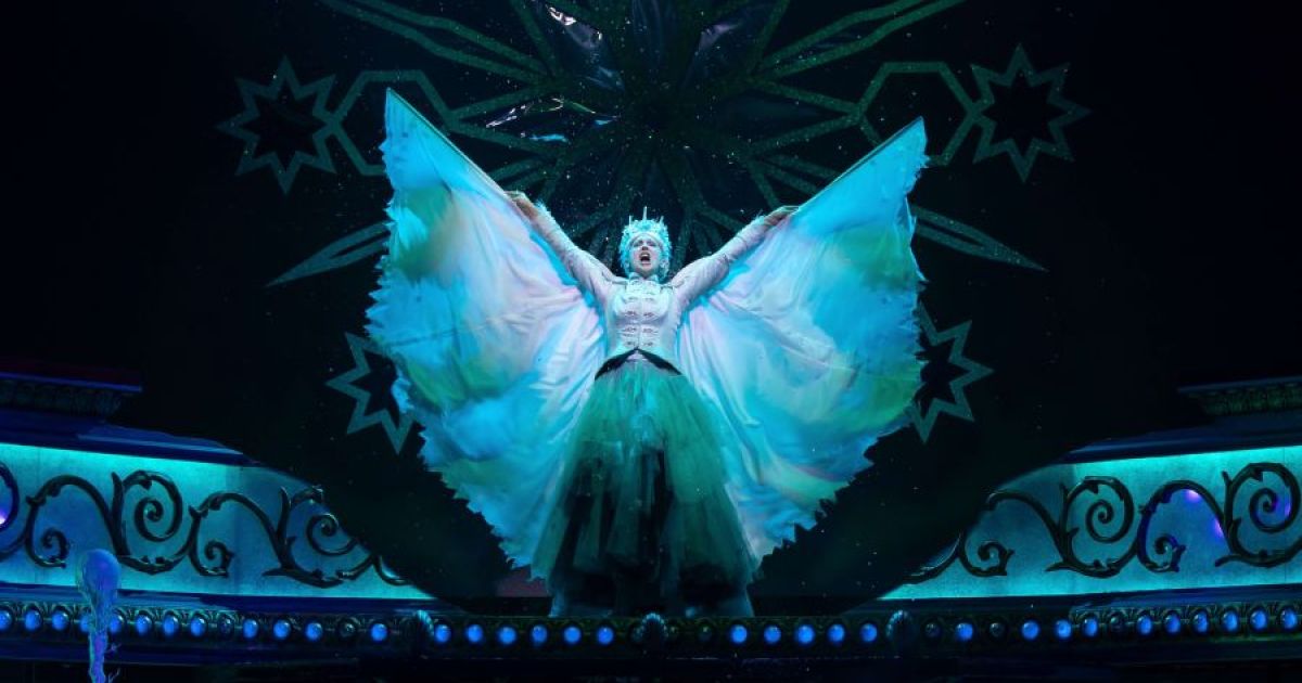 Claire Dargo as The Snow Queen, standing with her arms outstretched, reaching up to the sky. Black background with Snow Queen lit up in icy blue white.