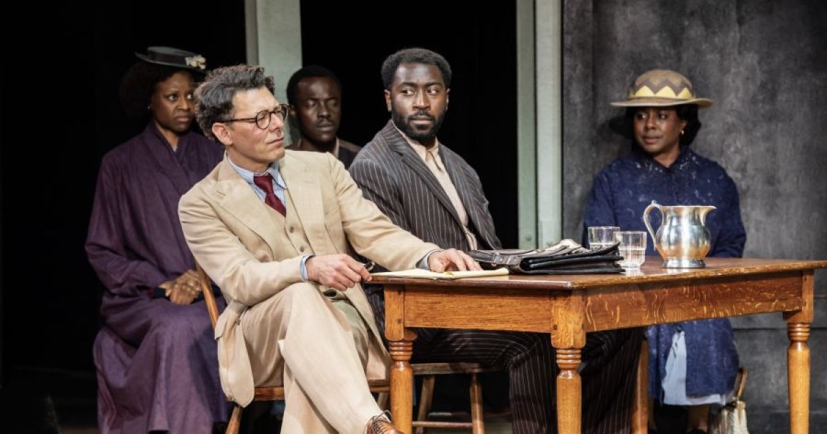 Court scene with Richard Coyle as Atticus Finch, Aaron Shosanya as Tom Robinson seated at a table.