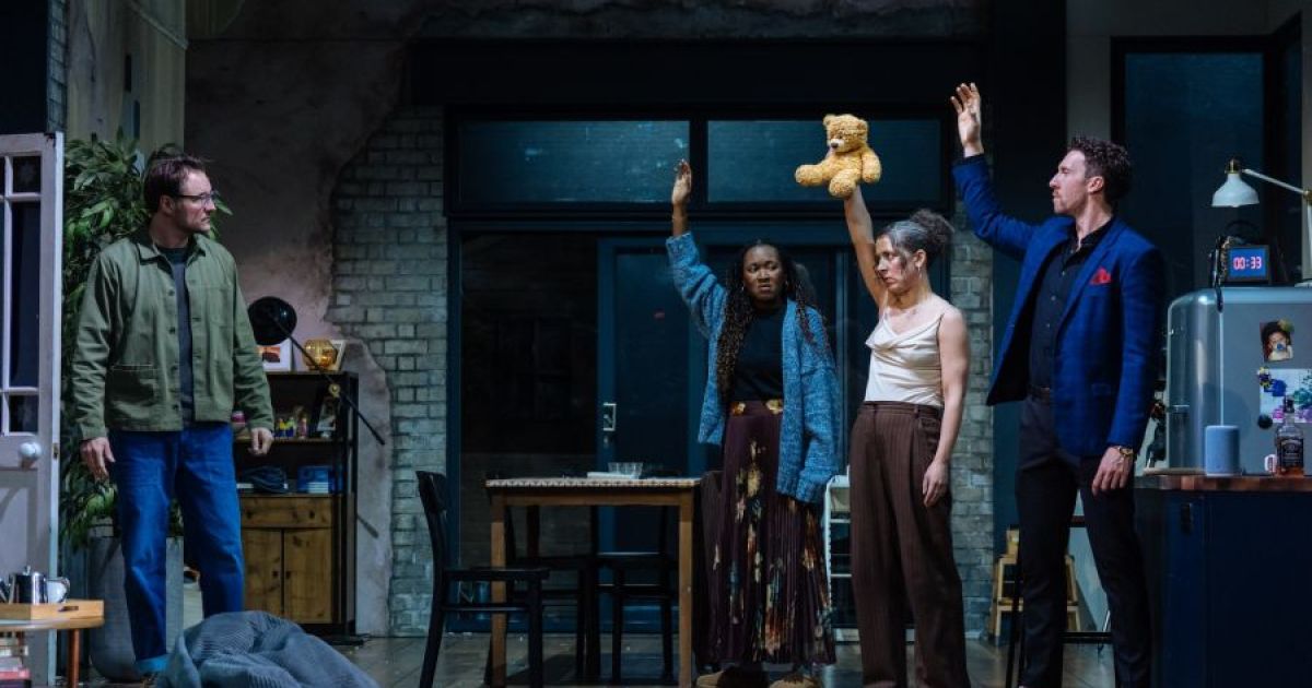 Four people stand in a room, three with their hands in the air, looking at the fourth,